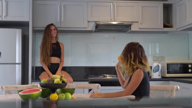 Two cheerful friends are in spacious kitchen, one long haired girl is on kitchen bench, the second is at a round table with vase of fruits on it, they have fun throwing an orange at each other