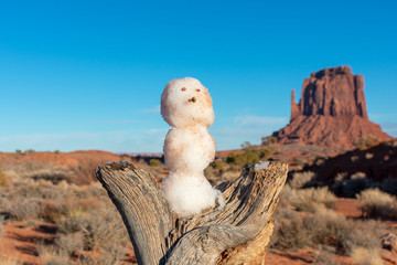 Close up. Small snowman on dead tree trunk under blue winter sky. Blurred desert landscape scenery with red sandstone butte in background.