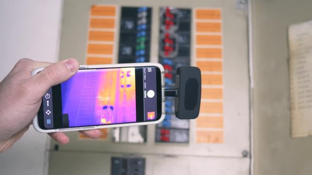 Electrician inspector using a thermal imaging camera attached to his smartphone as a tool to inspect the fuse box of the building.