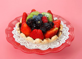 Fresh summer fruit tart with strawberries, kiwi, blueberries, boysenberries on a white doily on red plate pink background.