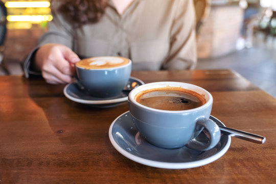 Closeup image of a hand holding a blue cup of hot black coffee with another latte coffee cup on wooden table in cafe