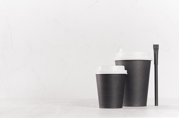 Coffee mockup - big and small black paper cups with white caps and sugar bag on white wood table with copy space, coffee shop interior.