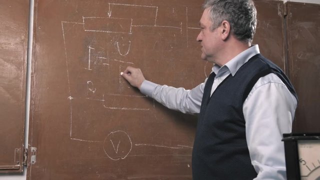 Senior teacher explains lesson, talking with students about physics and writing on blackboard. University gray haired professor giving lecture in science class.