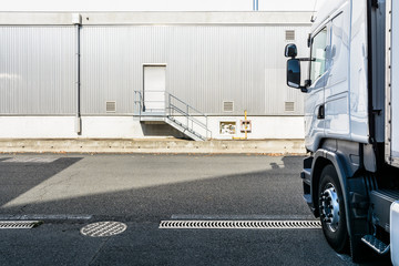 A white semitrailer truck parked opposite and perpendicularly to an industrial building with a pedestrian doorway at the top of a small staircase and covered with a grey corrugated iron siding. - obrazy, fototapety, plakaty