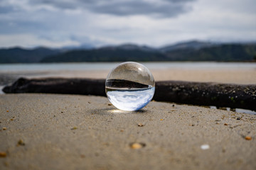 Fototapeta na wymiar lansball on the beach of Abel Tasman New Zealand, crystal glas ball on the beach with an old wood piece in the background, macro photography, beach and ocean background image New Zealand