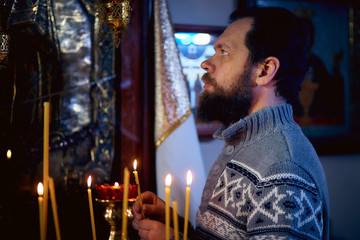 A Russian man with a beard stands in an Orthodox Church, lights a candle and prays in front of the icon.