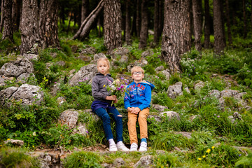 Boy with glasses and blue wear with bouquet of wild flowers and blonde girl near the forest in the summer mountains. Flowers for mother and sister for the woman’s day. RUSSIA, LAGO-NAKHI, July 2018