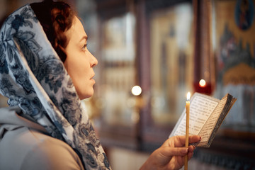 woman in the Russian Orthodox Church with red hair and a scarf on her head lights a candle and prays in front of the icon.