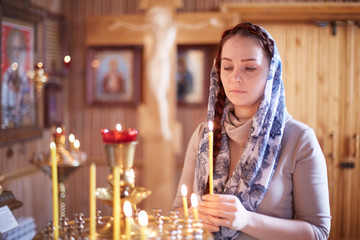 Russian beautiful Caucasian woman with red hair and a scarf on her head is in the Orthodox Church, lights a candle and prays in front of the icon.