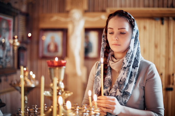 woman in the Russian Orthodox Church with red hair and a scarf on her head lights a candle and prays in front of the icon.