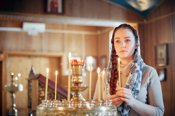 woman in the Russian Orthodox Church with red hair and a scarf on her head lights a candle and prays in front of the icon.