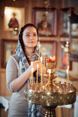 woman in the Russian Orthodox Church with red hair and a scarf on her head lights a candle and prays in front of the icon.