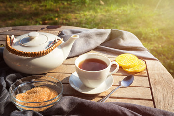 tea cup and a white teapot, brown sugar and lemon slices on a wooden garden table on a sunny morning, copy space