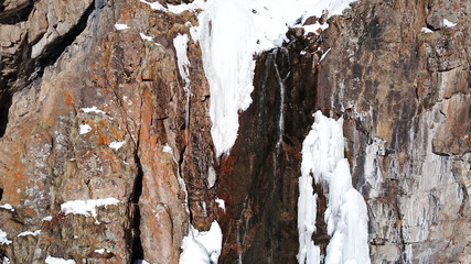 Freezing waterfall. Big icicles and snow rocks. Lots of ice on the rocks. Shooting with the drone. Water flows down the cliff. Snow cap, freezing river. Brown rocks.