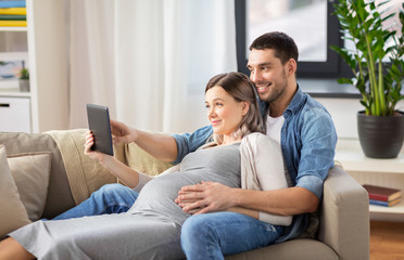 Obraz premium pregnancy, technology and people concept - happy man and his pregnant wife with tablet pc computer at home