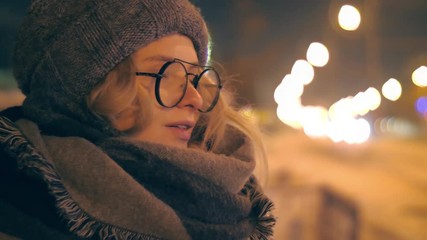 Beautiful young woman wearing fashion scarf standing in cold winter city street at night, looking at camera, closeup. Slow motion, shallow DOF, 4K UHD.