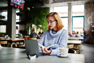 Cheerful young beautiful redhaired woman in glasses using her phone, touchpad and notebook while sitting at her working place on cafe with cup of coffee.