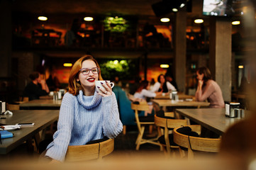Cheerful young beautiful redhaired woman in glasses sitting at her working place on cafe and drinking coffee.