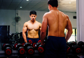 Fototapeta na wymiar An Asian man lifts a dumbbell in front of a mirror in a gym. Exercise in gym concept
