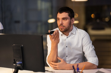 business, deadline and technology concept - businessman with computer using voice command recorder on smartphone at night office