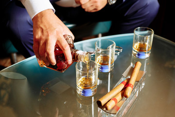 man pours alcohol in three glasses, three cigars lie in an ashtray