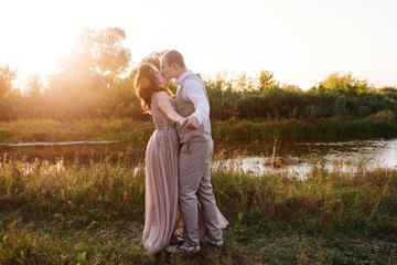 Naklejka na ściany i meble Wedding near the river in field at sunset with brown horse. Bride in light airy dress in color of dusty rose. Beige dress with sparkles. Light suit with bow tie. bride and groom embrace and kiss.