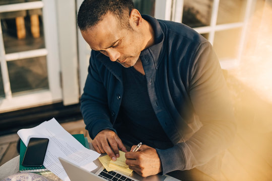 Mature man writing on adhesive note over laptop at table