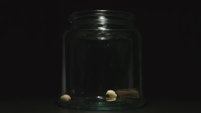 SLOW MOTION: Unpeeled peanuts fall into empty glass jar on black background - Front View