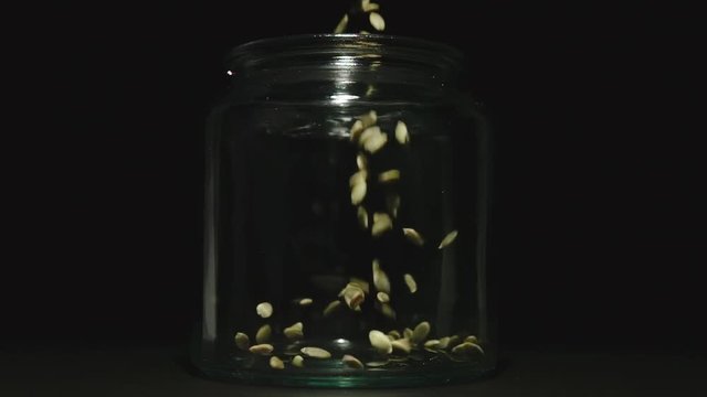 SLOW MOTION: Peeled pumpkin seeds fall into empty glass jar on black background - Front View