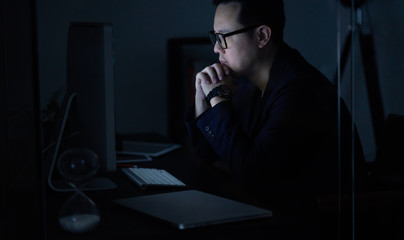 Young Asian businessman working on a laptop while watching computer at his office desk at night .