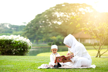 Muslim mother teaching her little son to reading the Quran on grass field near beautiful lake. Muslim family concept. 