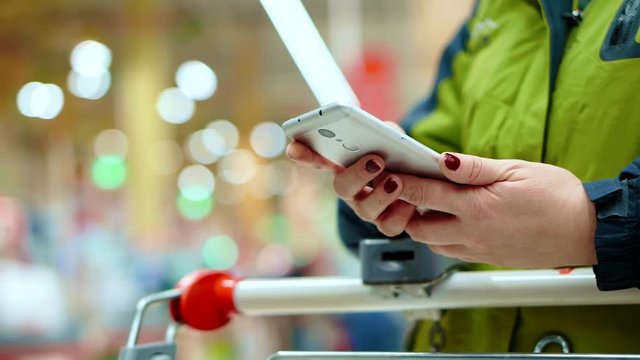 A woman in a supermarket holds a smartphone in her hand, she is leaning on a basket with goods, waiting for the queue at the checkout with products.