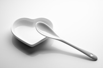 Ceramic white spoon is lying on a plate in the form of a heart on a clean background. Black and white picture with shadow. Concept love and food. Copy space. Template for mockup.