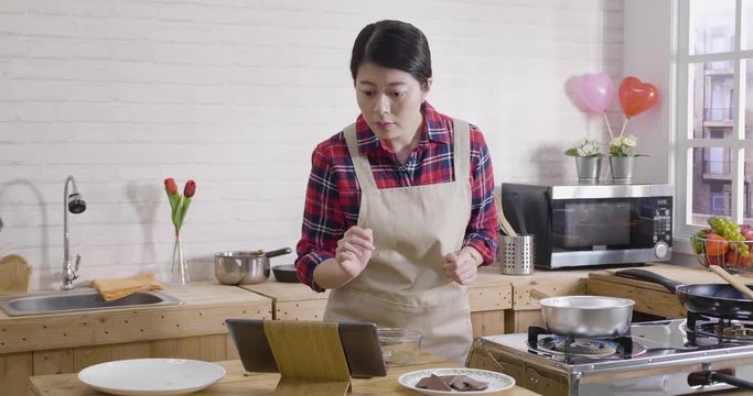 asian female turning on kitchen gas stove to boil water in wooden modern kitchen. young housewife making handmade chocolate surprise for husband. woman watch recipe on tablet peels cocoa into pieces.