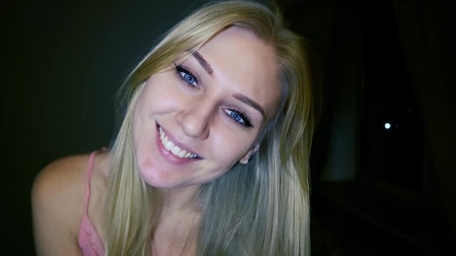 beautiful young blond woman with bright blue eyes sends an air kiss to the camera and smiles, having bent her head, dark background, slow motion