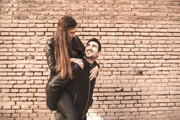 Young girl climb on the back of her boyfriend, piggyback, in front of a brick wall having fun and laughing loudly.