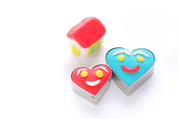 横長の白バックに置かれた粘土で作ったハート型の笑顔の男女と粘土で作った小さな家