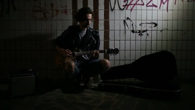 A street musician playing on guitar in the underpass. Vagrant lifestyle. Playing in the underpass to make a living. Unemployed musician. Future rock star. Video.