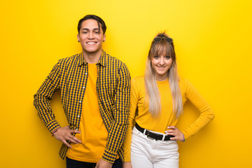 Young couple over vibrant yellow background posing with arms at hip and smiling