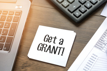 Desk with business accessories, calculator, documents with charts, fountain pen, laptop and paper for notes with text Get a grant