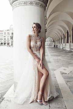 Beautiful bride in elegant, luxury ivory dress and messy updo hairstyle, posing near column in Venice, Italy