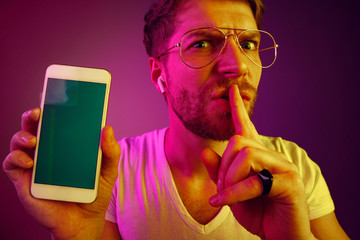 Indoor portrait of attractive young man isolated on pink neon background, holding blank smartphone, smiling at camera, showing screen, feeling happy and surprised. Human emotions, facial expression