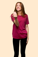 Young redhead girl inviting to come with hand. Happy that you came on isolated yellow background