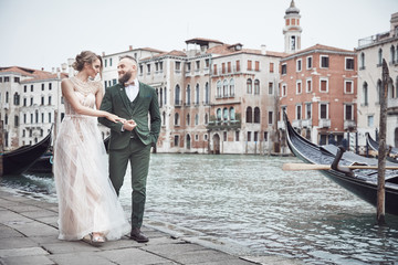 Fototapeta na wymiar Amazing wedding couple walk near the canal in Venice. Elegant woman in luxury ivory dress, messy updo hair, man in green three-piece suit
