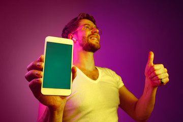 Indoor portrait of attractive young man isolated on pink neon background, holding blank smartphone, smiling at camera, showing screen, feeling happy and surprised. Human emotions, facial expression