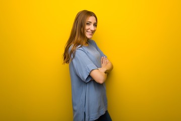 Obraz premium Young redhead girl over yellow wall background keeping the arms crossed in lateral position while smiling