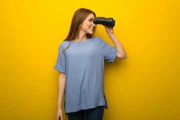 Young redhead girl over yellow wall background and looking in the distance with binoculars
