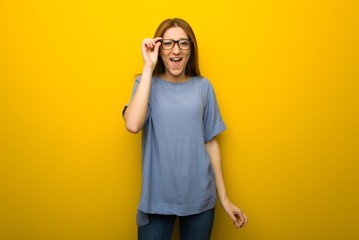 Young redhead girl over yellow wall background with glasses and surprised