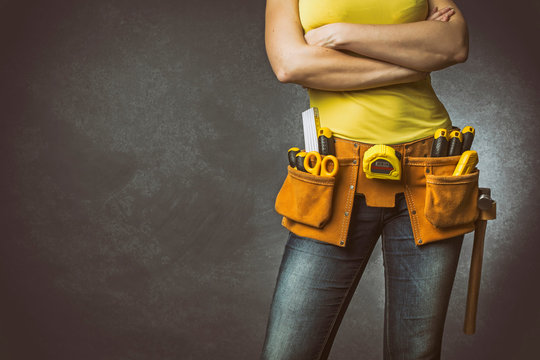 Handywoman with folded arms and tool belt on concrete background.