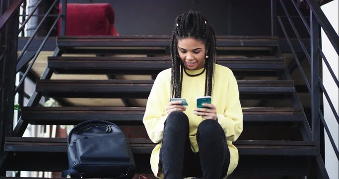 Closeup beautiful multi ethnic lady with a long dreadlocks hair sitting on modern stairs and using her smartphone and bank card order something online or check her internet backing card. 4k
